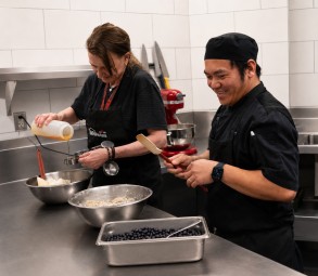 Two 2G2B Healthy team members in the prep kitchen, one measuring ingredients into mixing bowls and the other noting a recipe